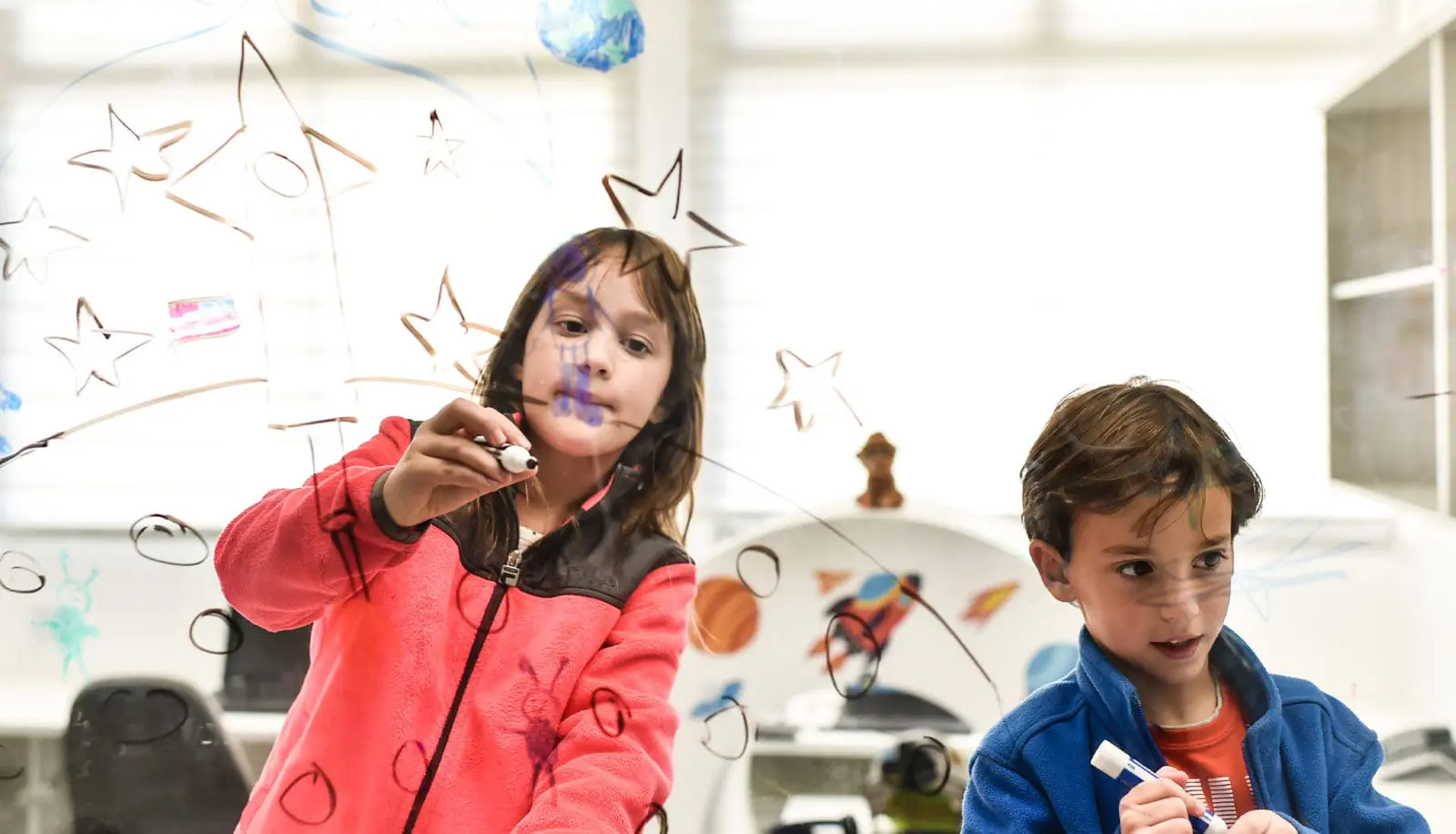 Young girl and boy drawing stars and a rocket on a clear glass window