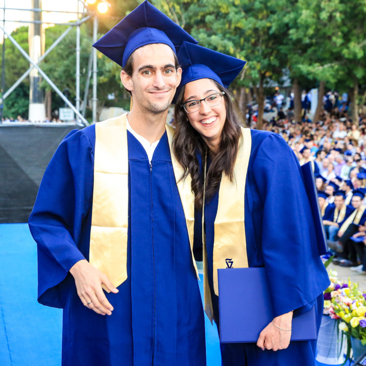 A posed photo of a male and female Technion student at graduation, in cap and gown with diplomas.