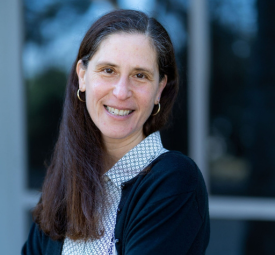 A headshot of a woman with long brown hair, a light colored shirt and dark sweater.