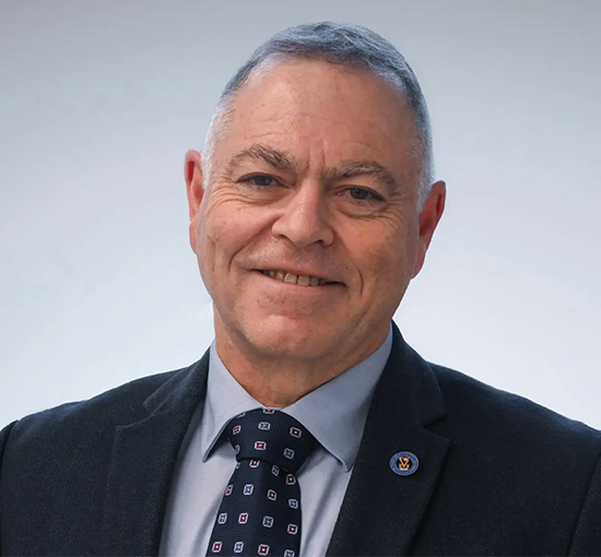 Rafi Aviram headshot from chest height. Suit and tie visibile, gray background