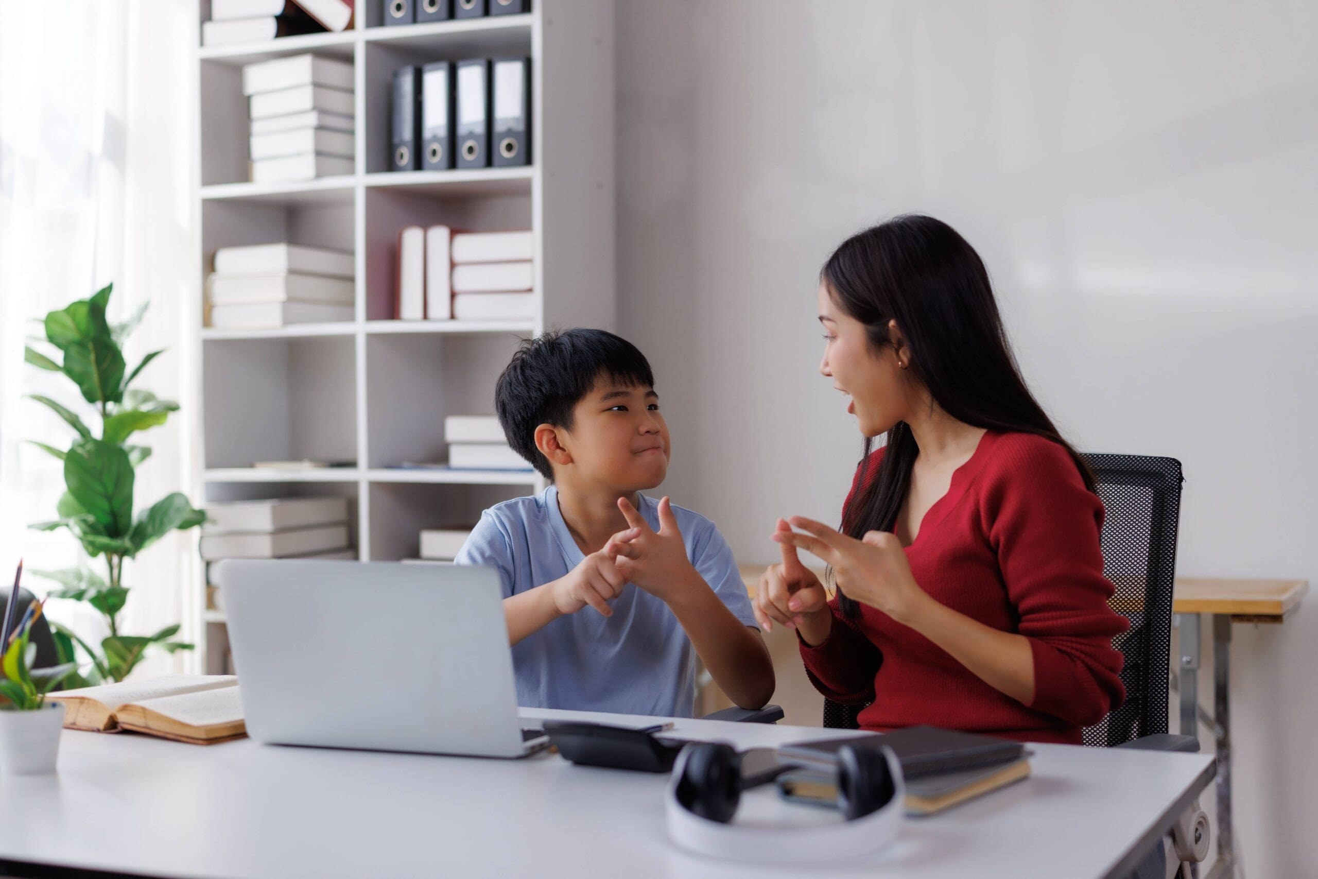 Young boy student in blue, seated left, and young woman teacher in red, seated right, learning sign language in a home office setting