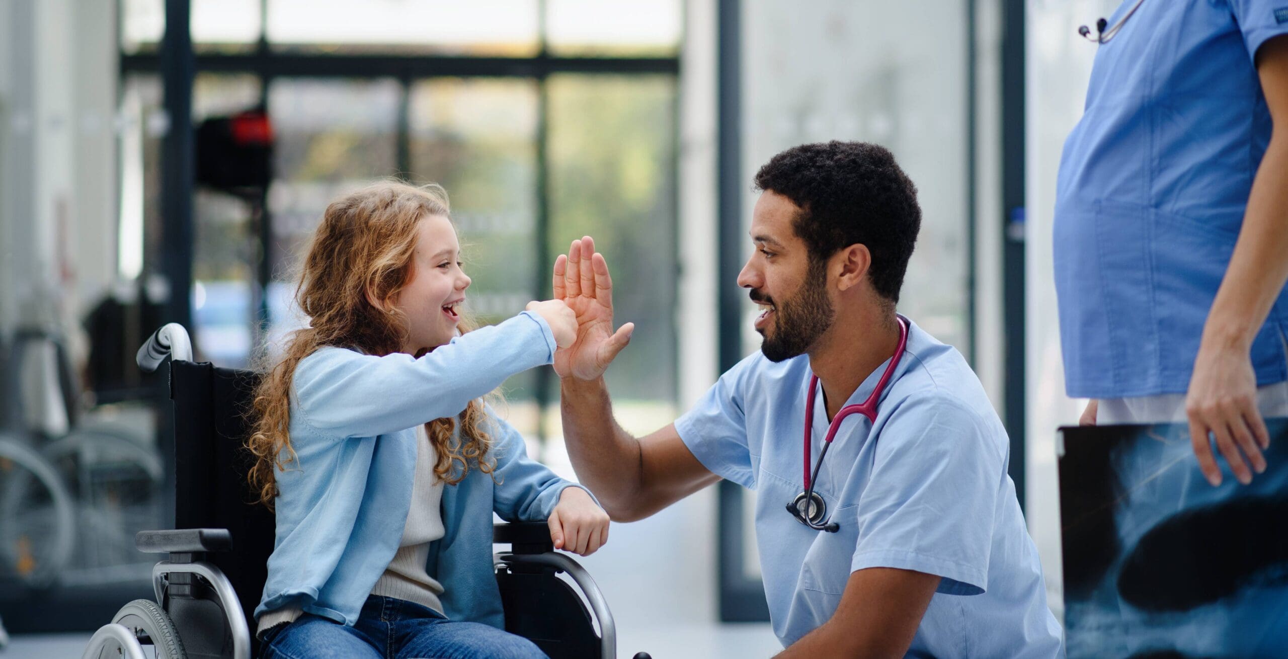 Young girl in wheelchair, left, high fives kneeling doctor, right, in hospital setting.