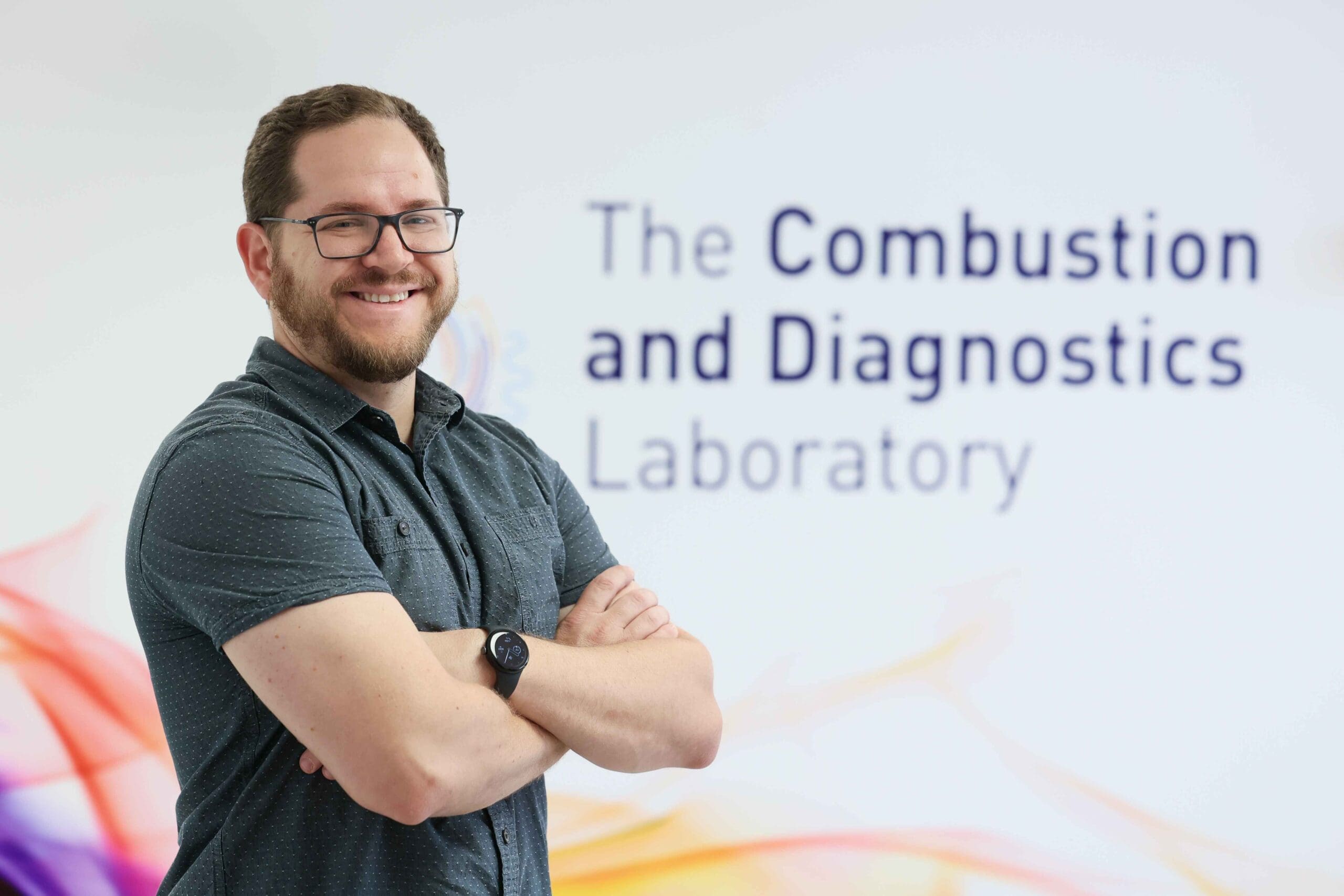 Professor Joe Lefkowitz standing to the left, arms crossed, in front of a sign for The Combustion and Diagnostics Laboratory.